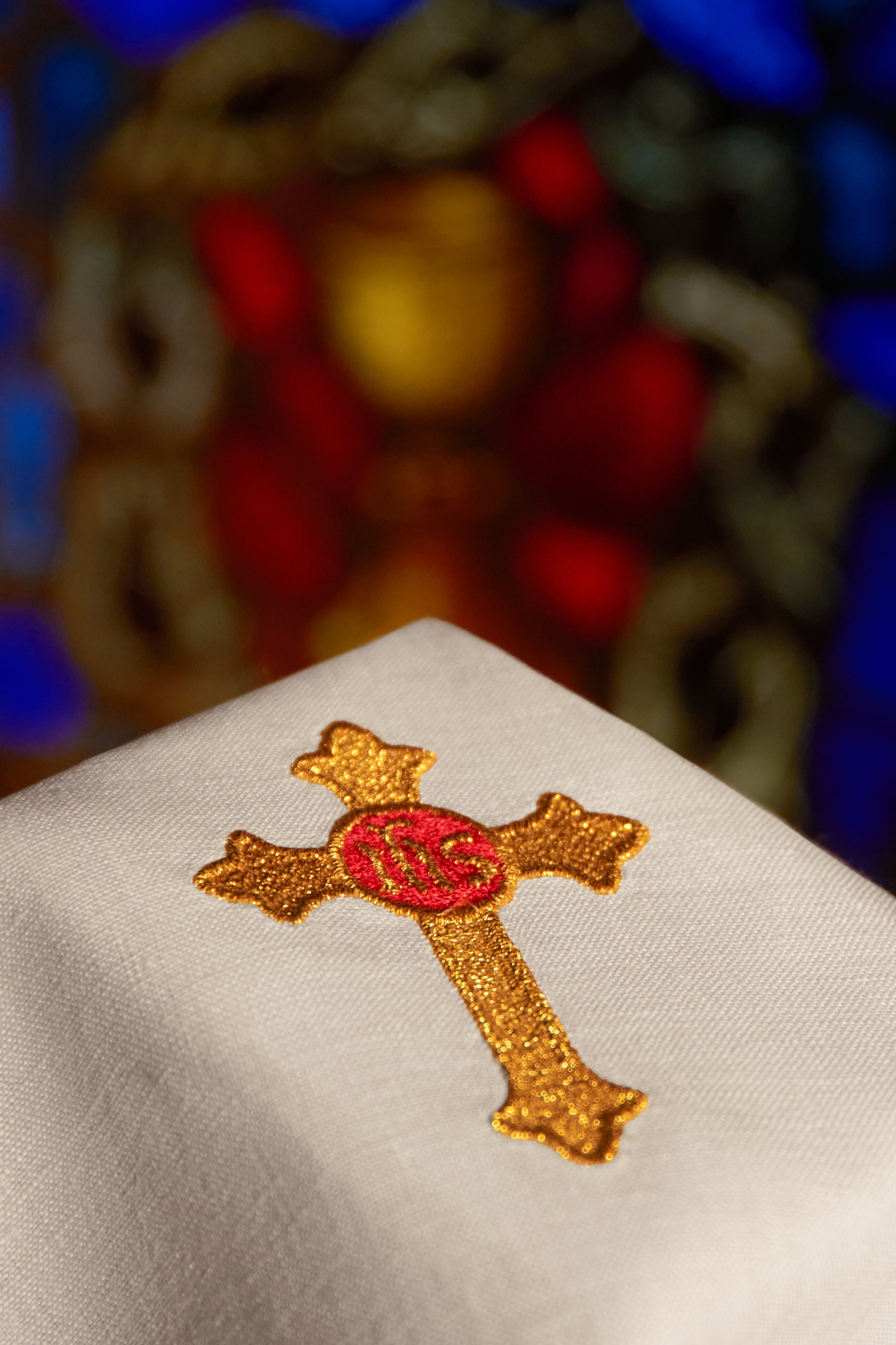 A close-up of a white fabric cross embroidered with gold thread and a red center, set against a blurred multicolored stained-glass background.